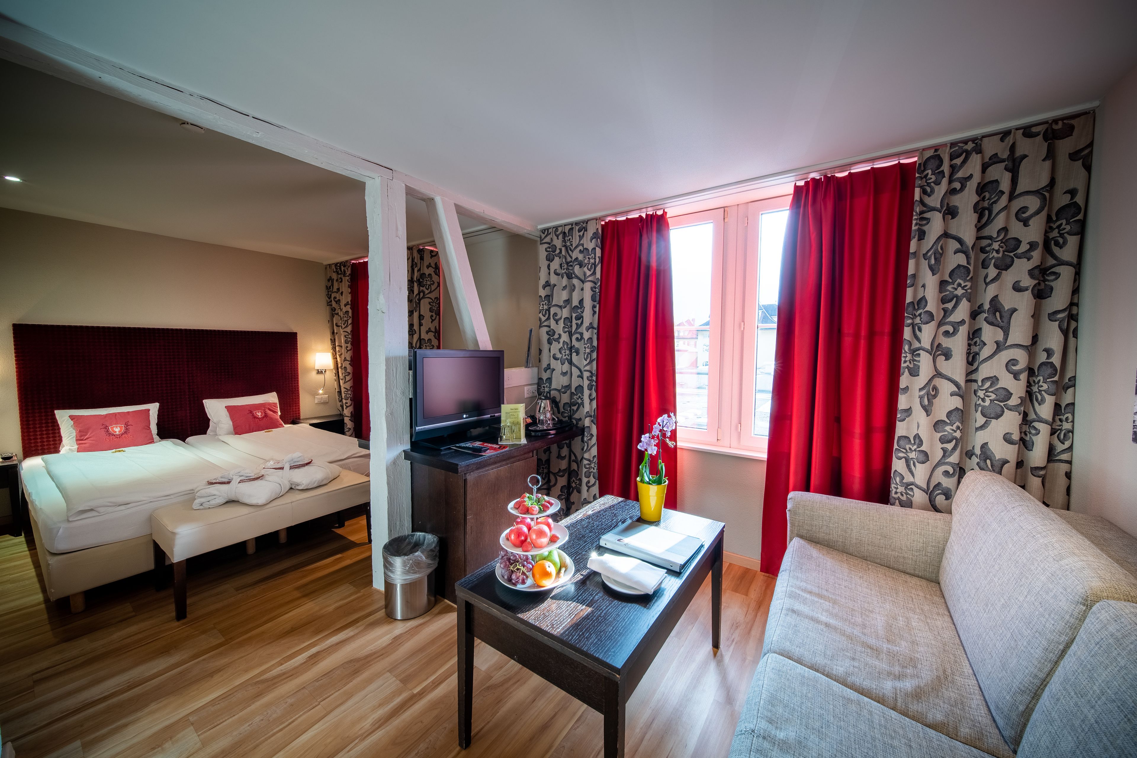 Een hotelkamer met een bed, een televisie, een klein tafeltje met fruit, een bank en een raam met rode gordijnen.