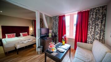 Een hotelkamer met een bed, een televisie, een klein tafeltje met fruit, een bank en een raam met rode gordijnen.