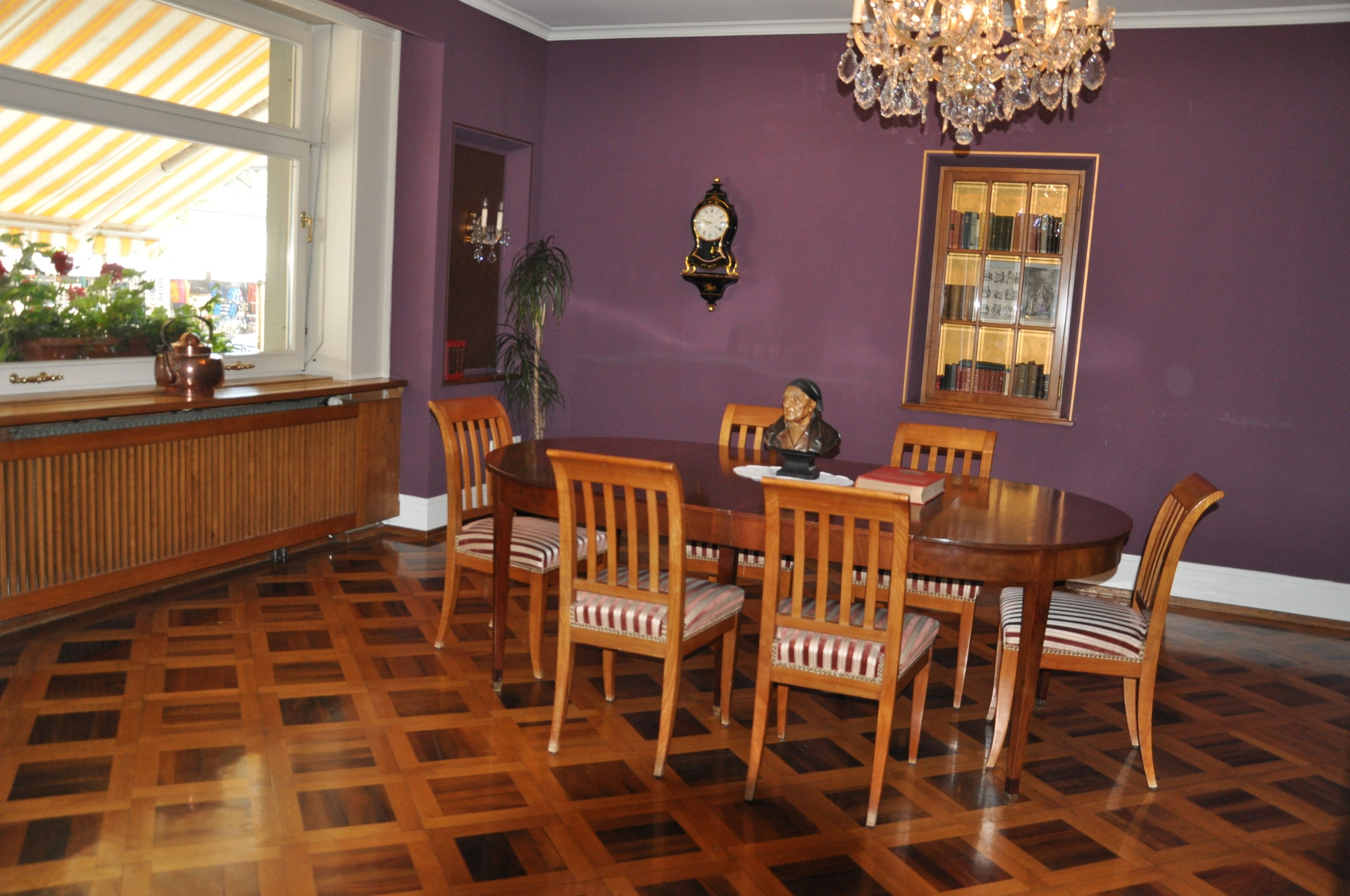 Un comedor con una mesa y sillas de madera, una araña, un reloj y una ventana con vistas a plantas.