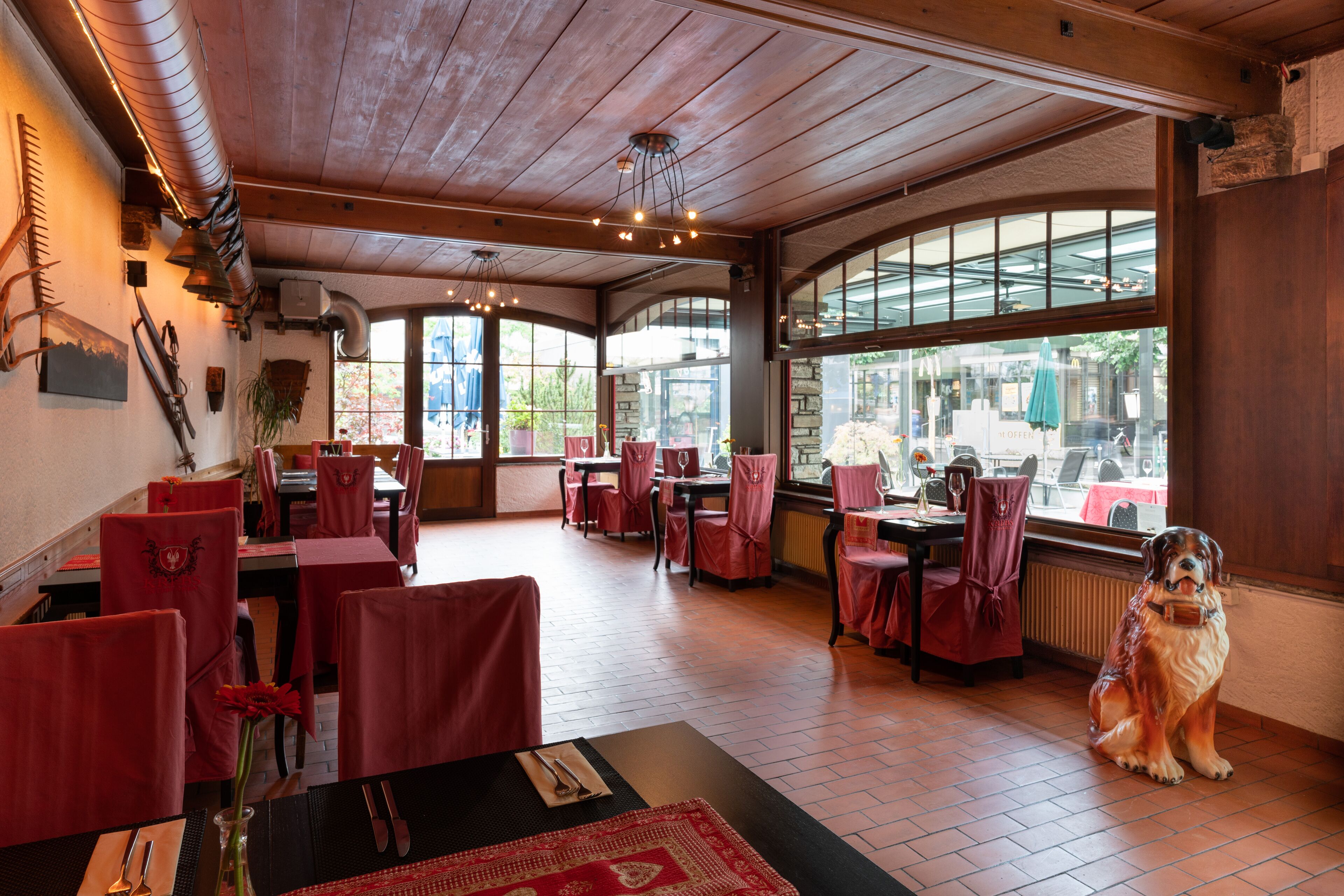 A restaurant with wooden ceilings and large windows. Tables are set with red tablecloths and chairs. A dog is sitting by the window.