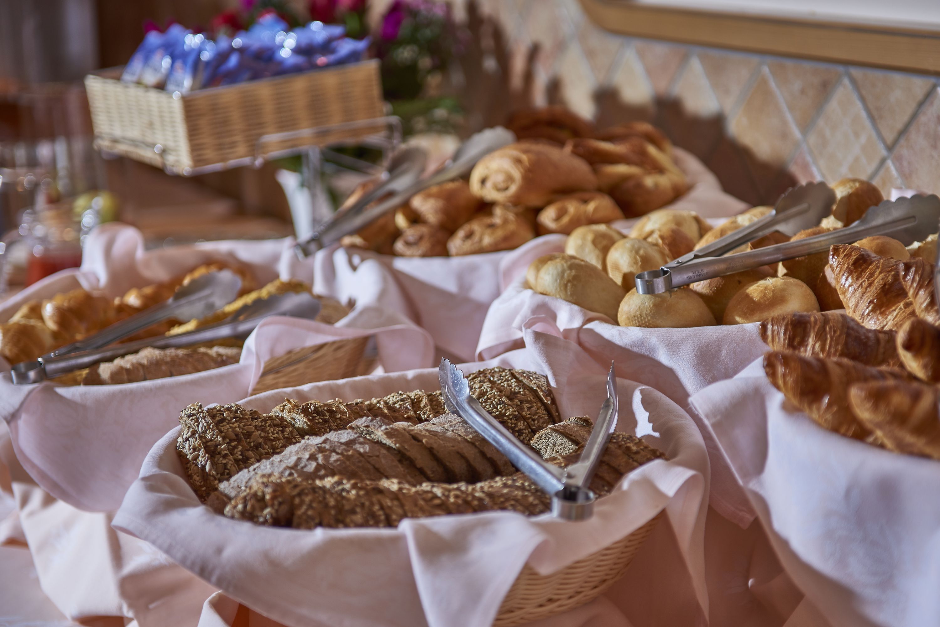 A buffet with various breads and pastries, including croissants and a loaf of bread, displayed in baskets with tongs.