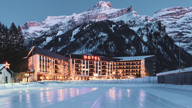 Un hôtel situé au pied d’une montagne enneigée, avec un grand bâtiment à plusieurs étages et une enseigne « HÔTEL » bien visible.