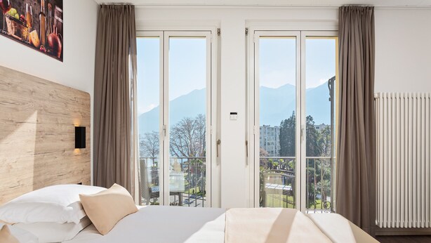 A hotel room with a bed, a wooden headboard, a large window with curtains, and a view of mountains. - Hotel DU LAC Locarno (Locarno)