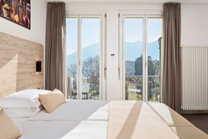 A hotel room with a bed, a wooden headboard, a large window with curtains, and a view of mountains. - Hotel DU LAC Locarno (Locarno)