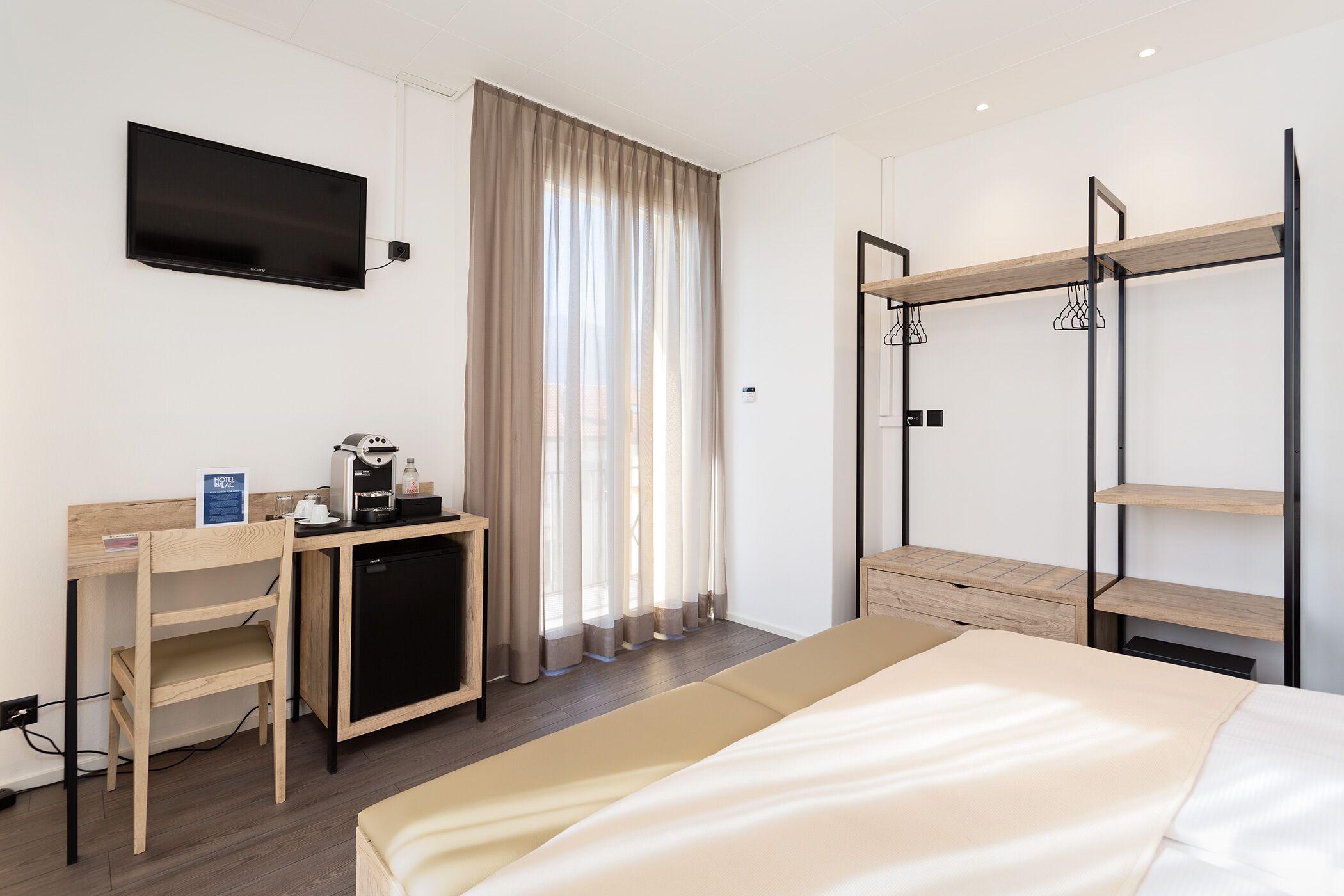 A hotel room with a bed, a desk with a coffee machine, a TV mounted on the wall, and a window with curtains.