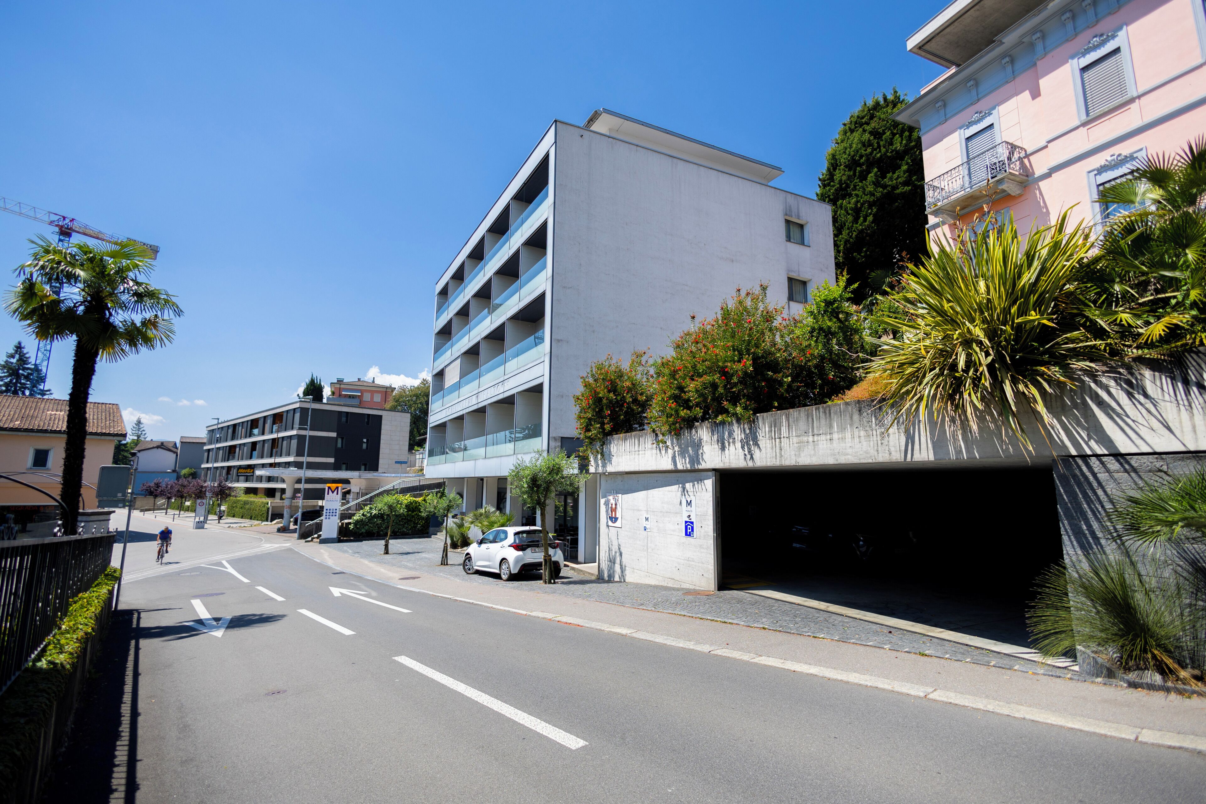 A multi-story building with a parking garage entrance, a cyclist on the road, and a clear blue sky.