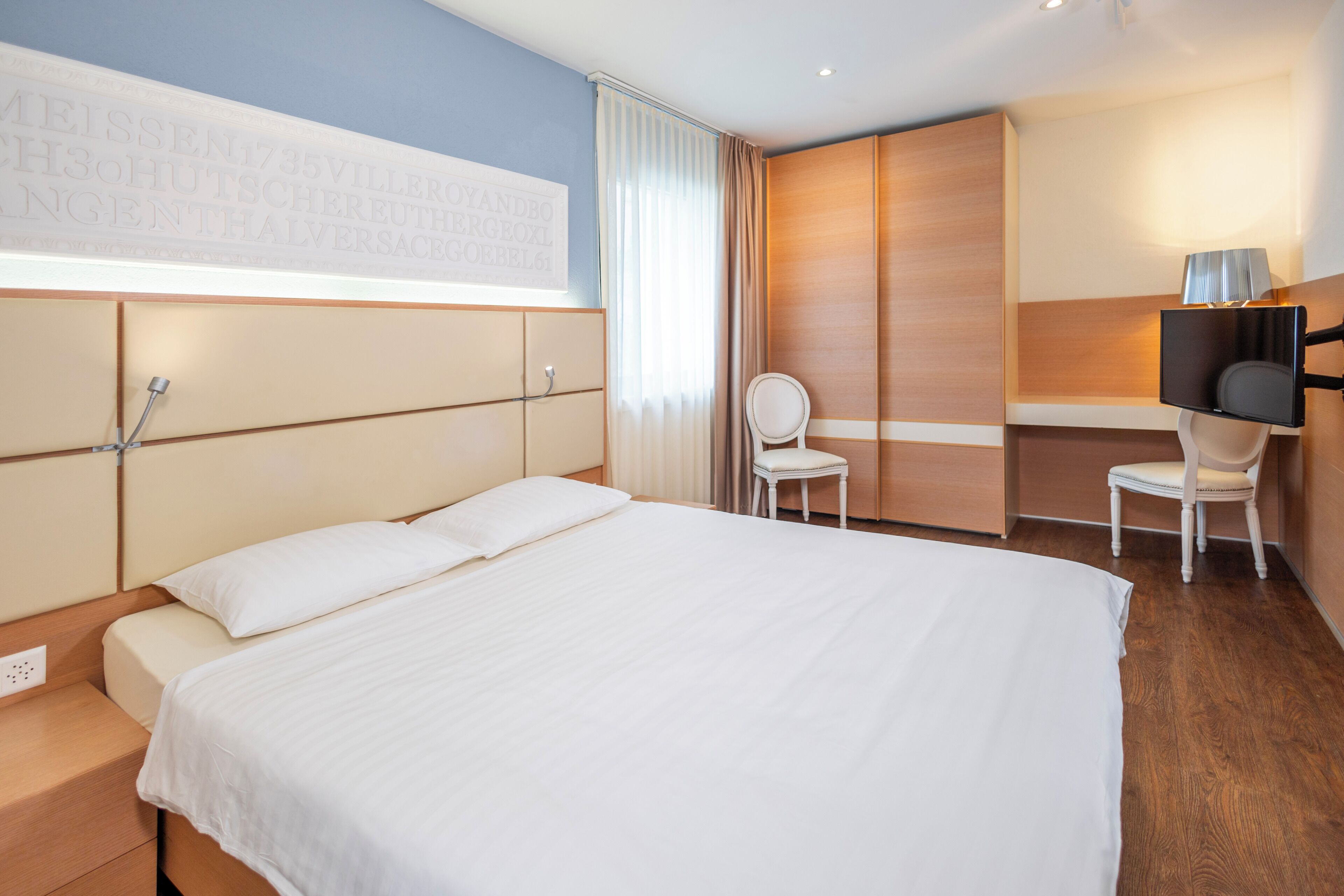 A hotel room with a bed, a chair, a TV, and a large window with curtains.