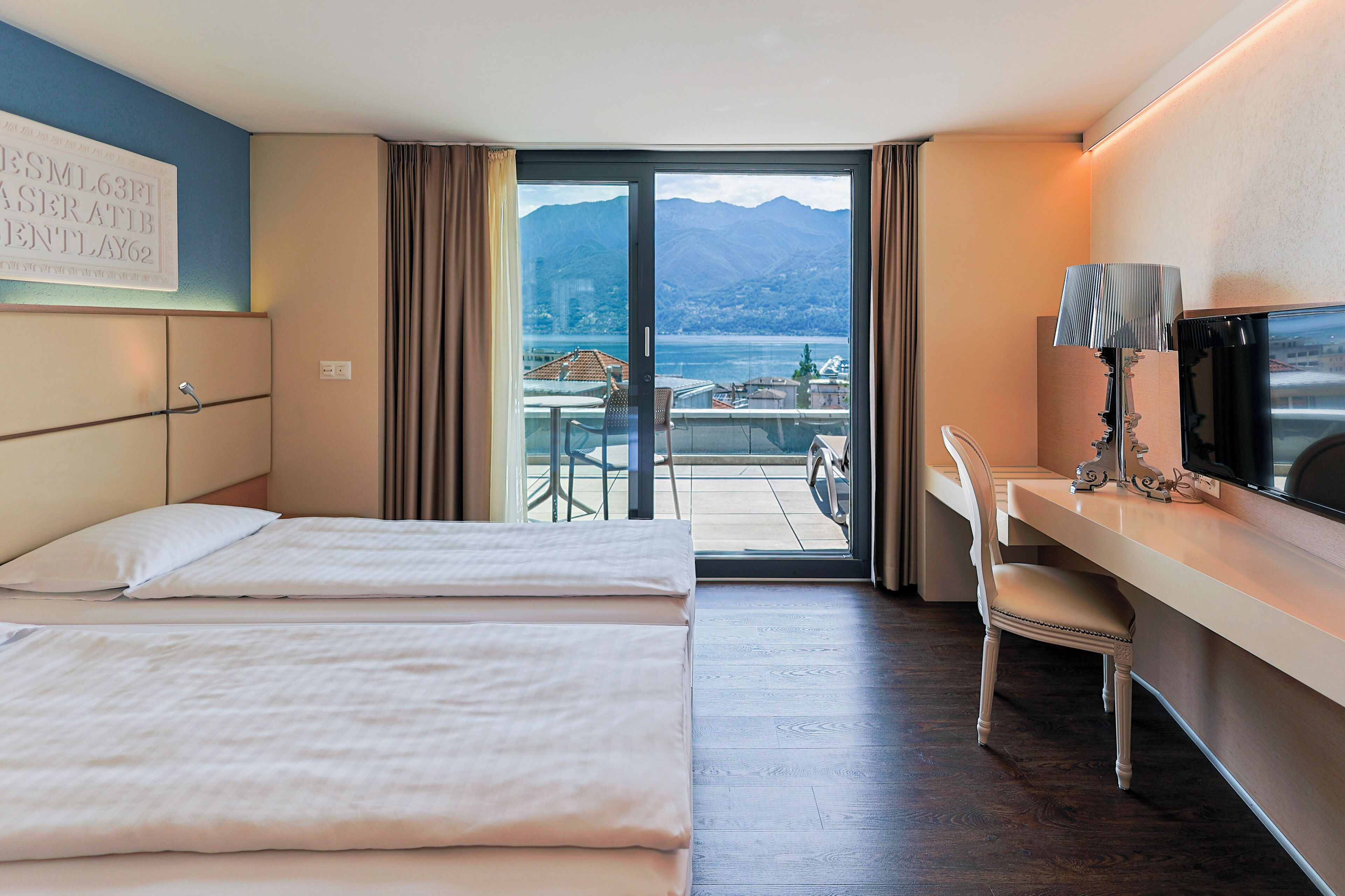 Una camera d'albergo con due letti, una scrivania con una sedia, una televisione e un balcone con vista su montagne e un lago.