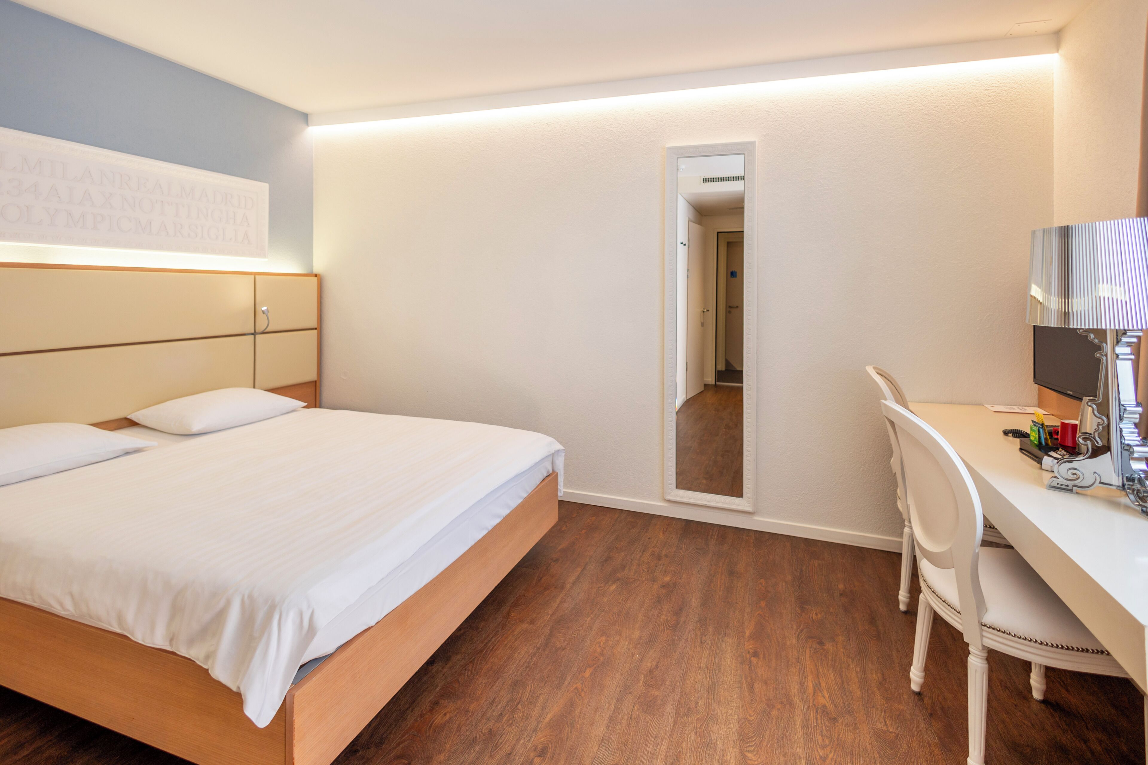 A hotel room with a bed, a desk, a chair, a mirror, and a wall with text.