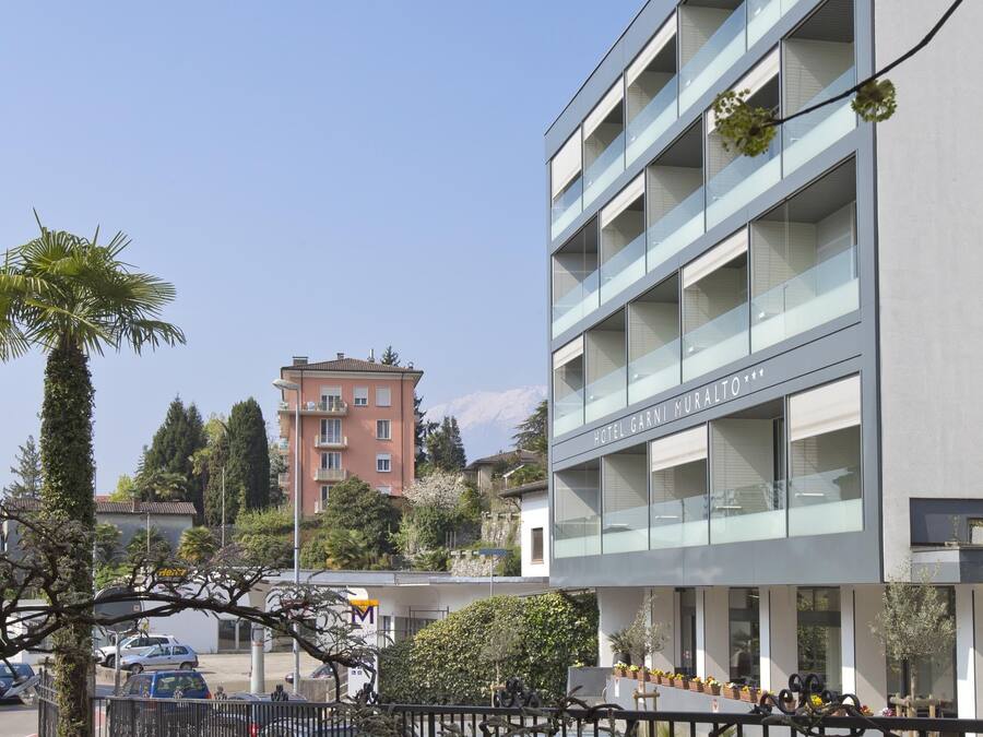 Um hotel moderno com fachada de vidro, várias varandas e estacionamento com carros.