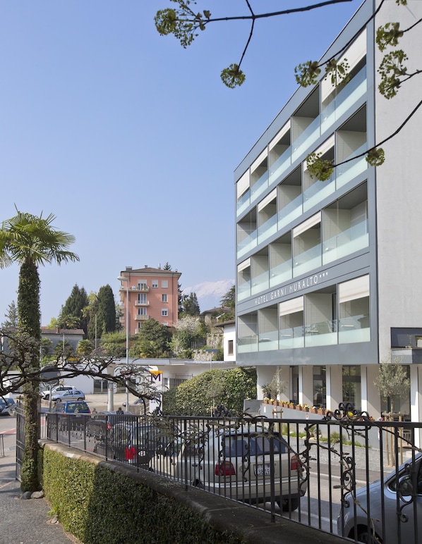 A modern hotel with a glass facade, multiple balconies, and a parking area with cars.