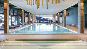 Una piscina interior moderna con paredes de vidrio transparente, vigas de madera y zonas de estar.