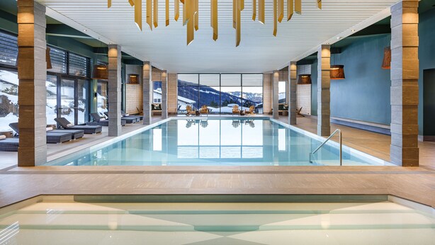 A modern indoor swimming pool with clear glass walls, wooden beams, and seating areas.