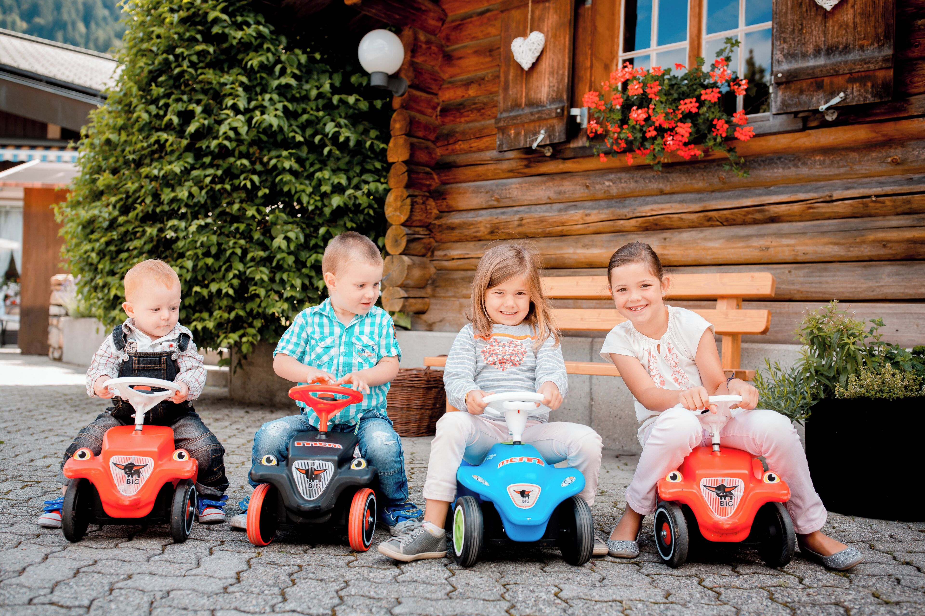 Three children riding toy cars in front of a wooden cabin with a bench and potted plants.