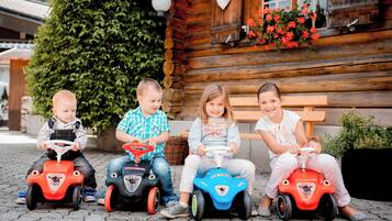 Three children riding toy cars in front of a wooden cabin with a bench and potted plants.
