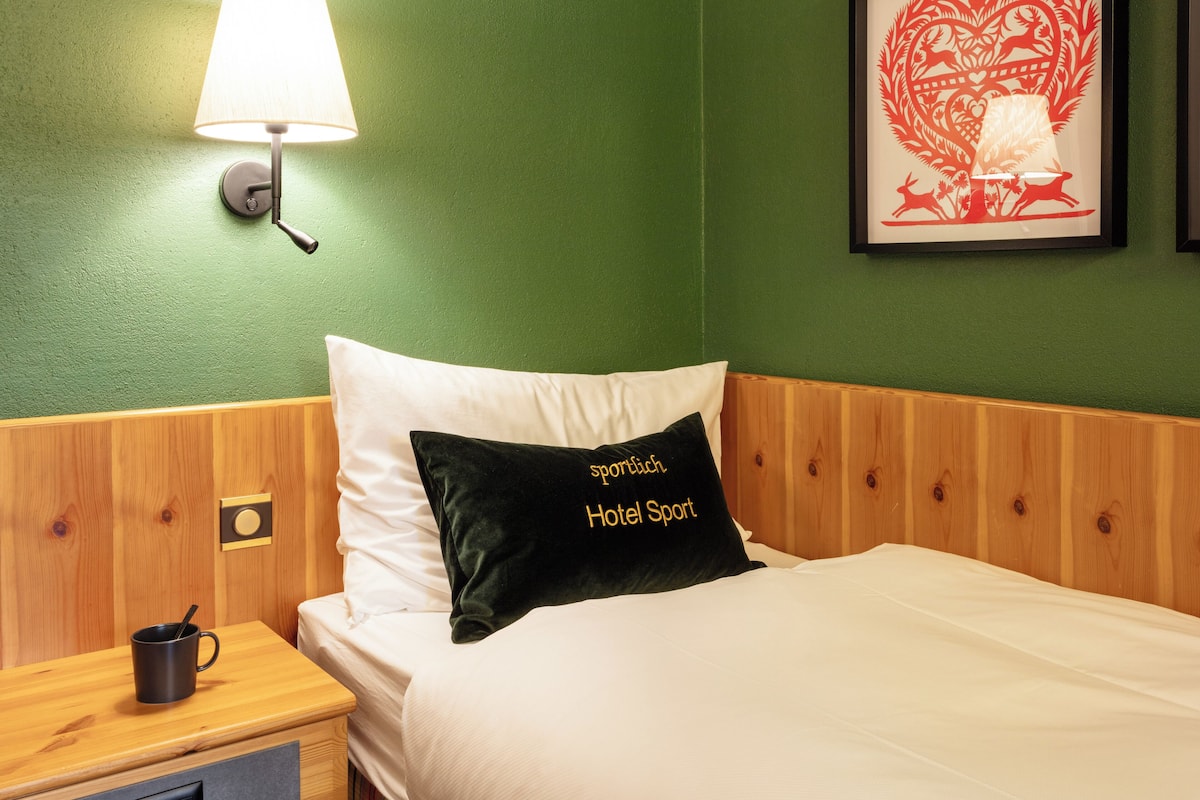 A hotel room with a green wall, a wooden headboard, a white bed, a black pillow with "Hotel Sport" embroidered, a bedside table with a black mug, and a wall-mounted lamp.
