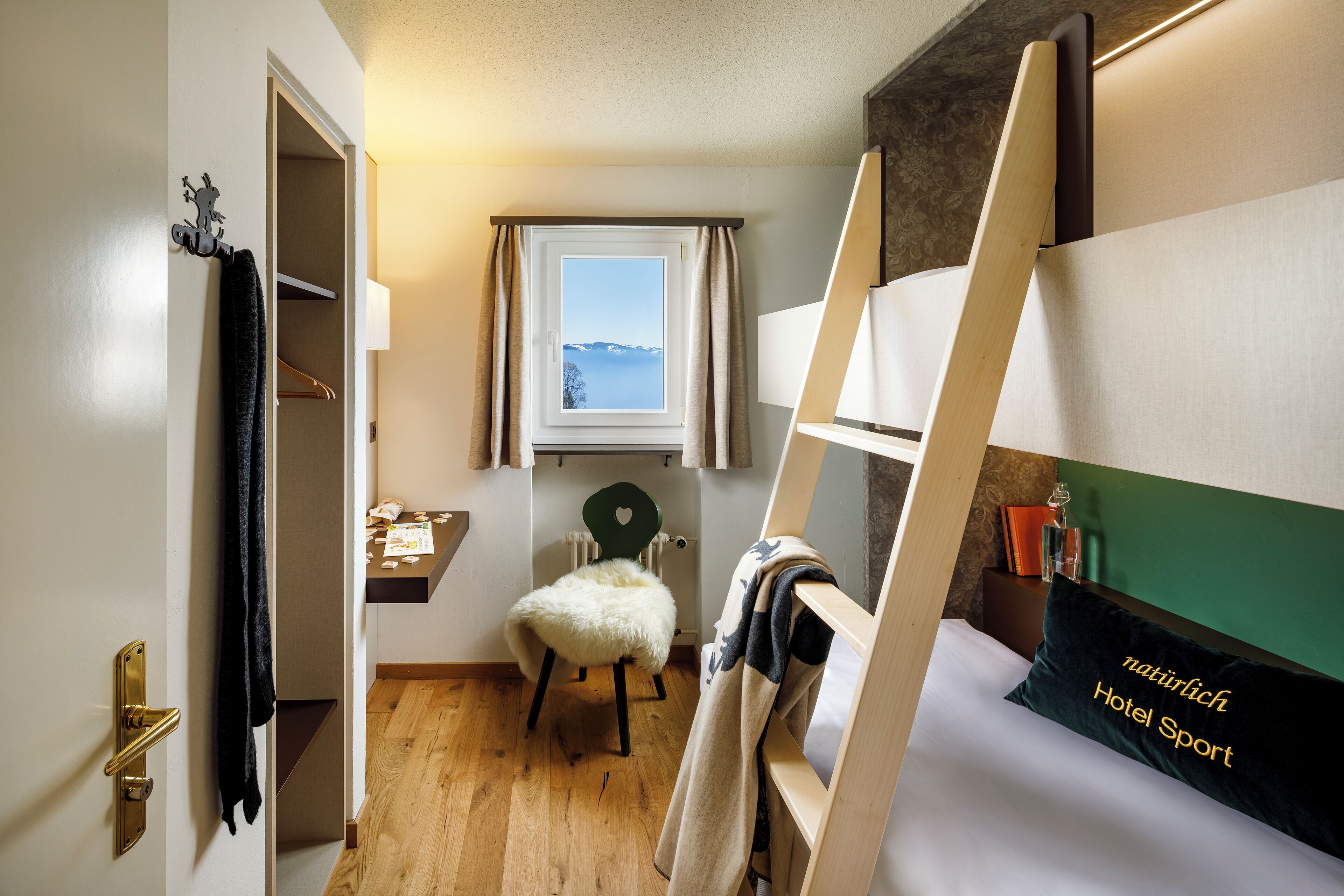 A compact hotel room with a bunk bed, a small desk with a lamp, a chair, and a window with a view of the mountains.