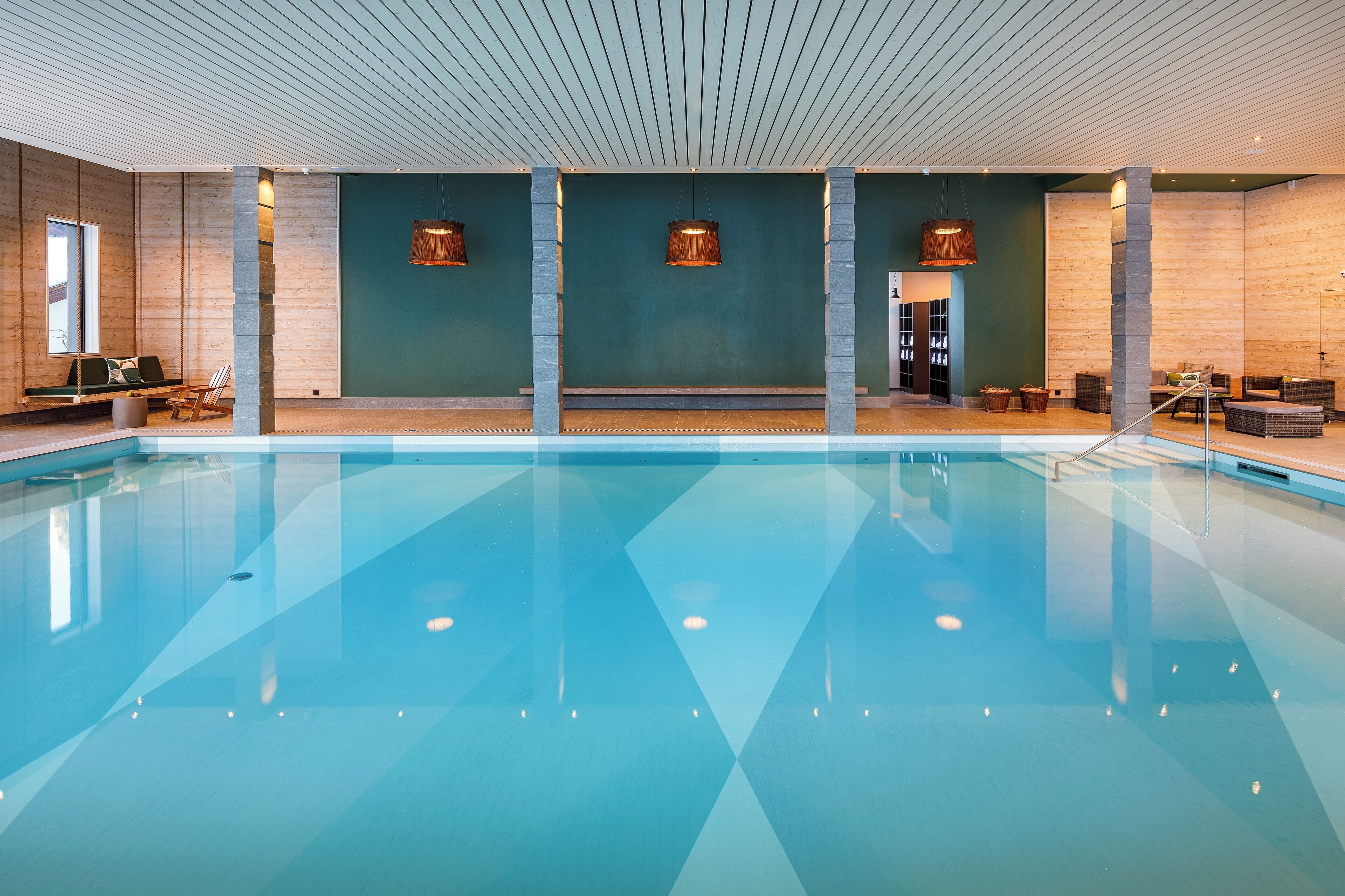 Ein großes Hallenbad mit klarem, blauem Wasser, Holzverkleidung und Sitzbereichen mit Kissen und Körben.