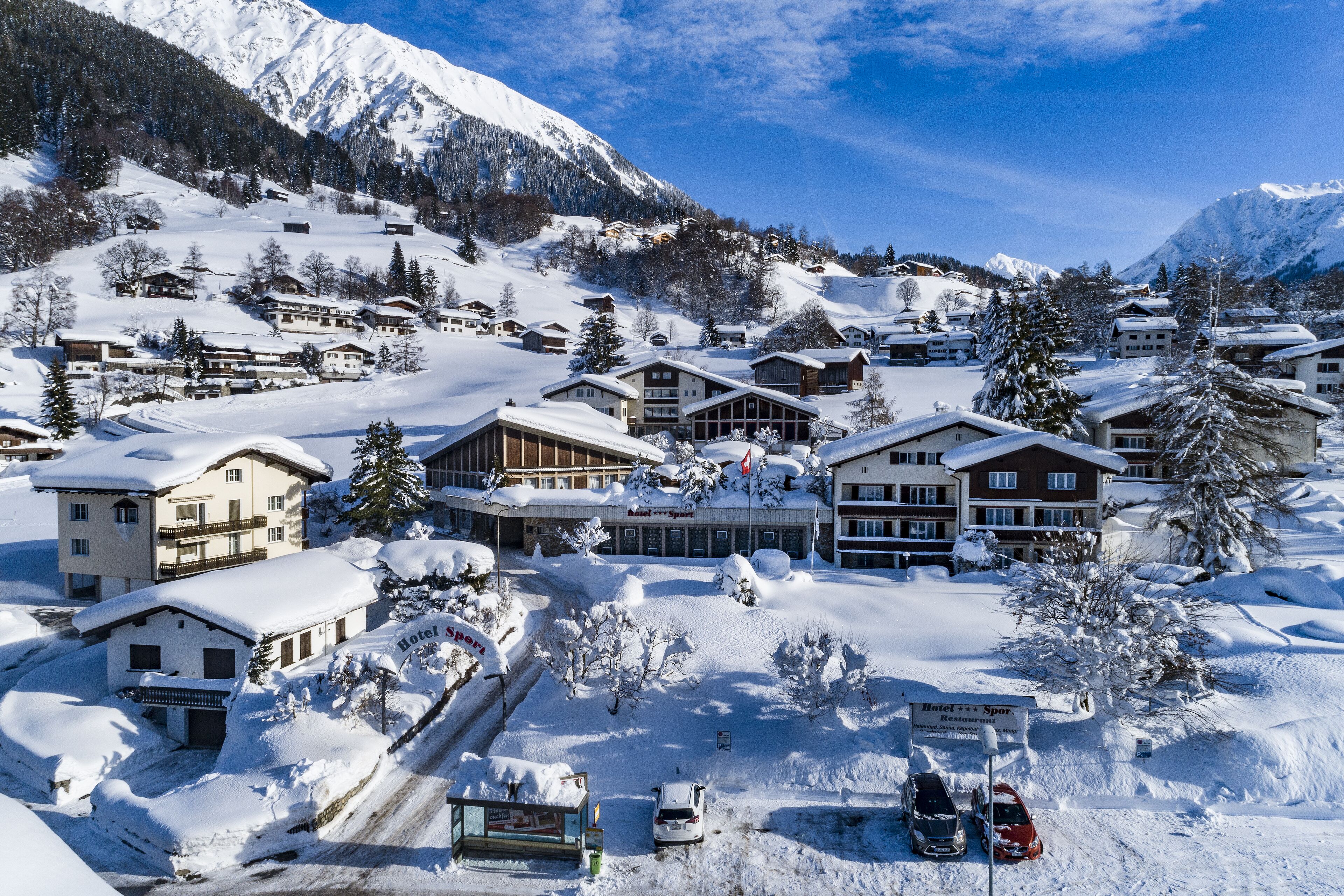 Uma aldeia montanhosa coberta de neve, com edifícios em estilo chalé, uma estância de esqui e um hotel chamado "Hotel Sport".