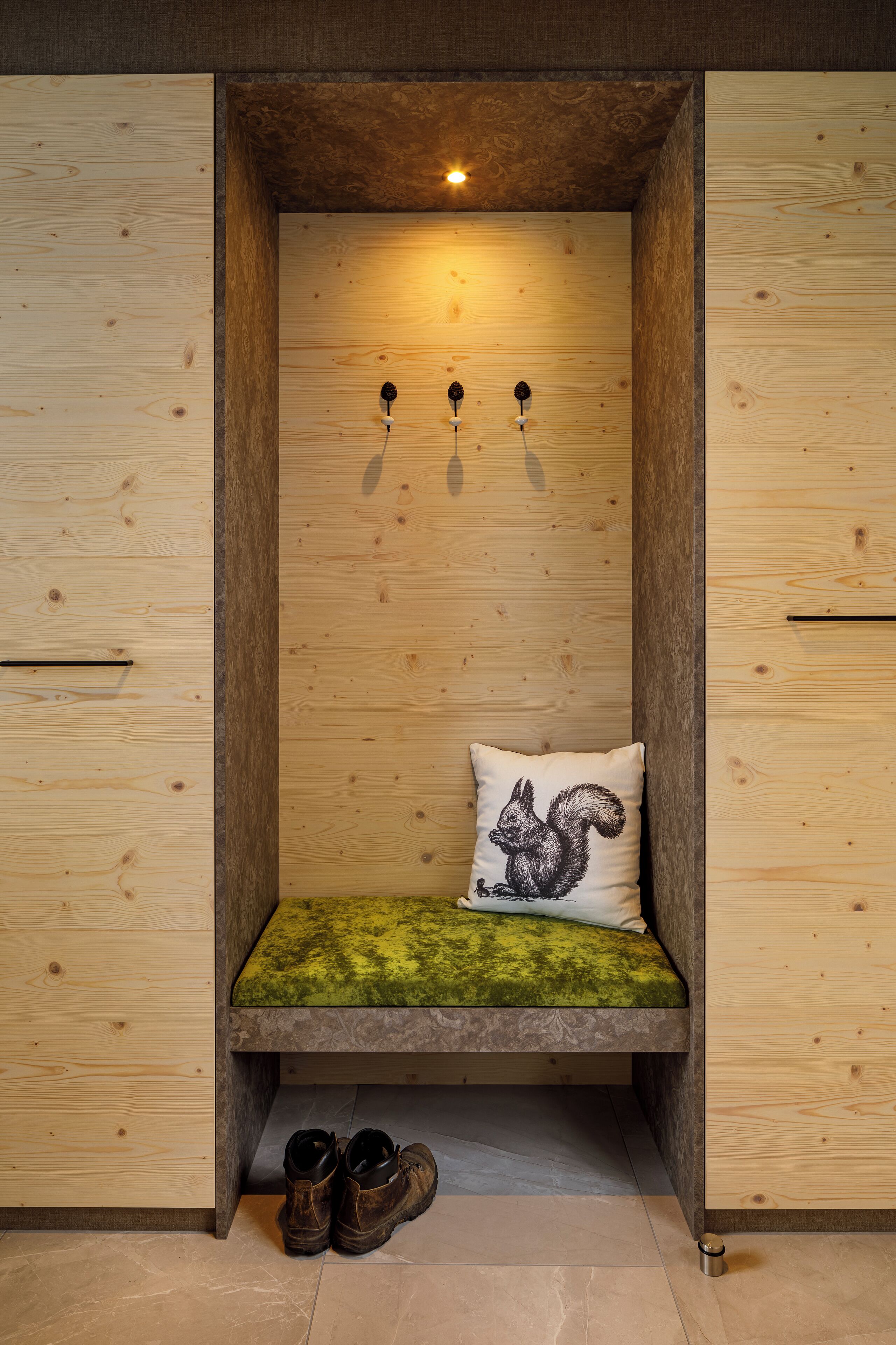 A wooden bench with a green cushion and a pillow with a squirrel design, under a slanted ceiling with three wall-mounted lights.