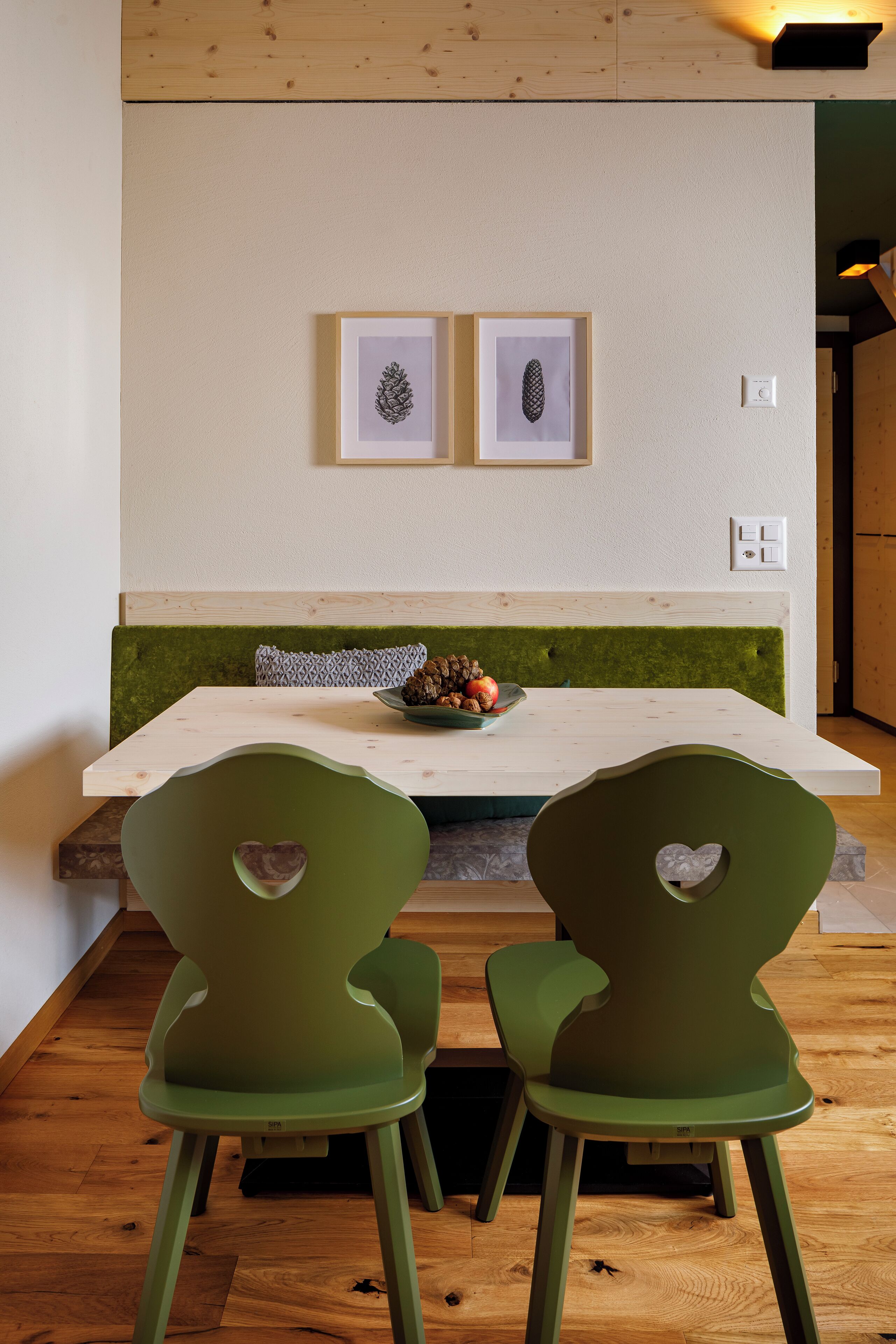 Un comedor con una mesa de madera, dos sillas verdes y un cuenco de fruta. Hay dos cuadros en la pared sobre la mesa.