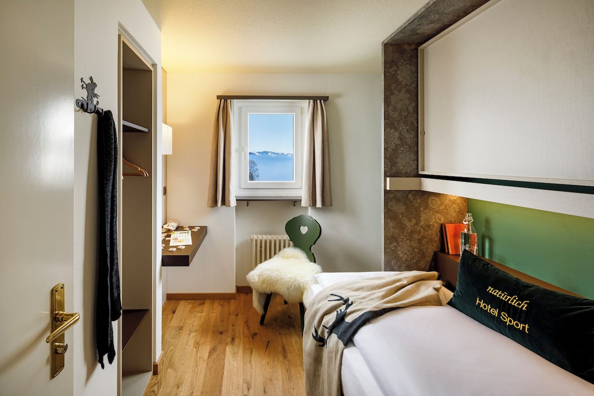 A hotel room with a bed, a chair, a window with a view, a radiator, and a shelf with books and a bottle.