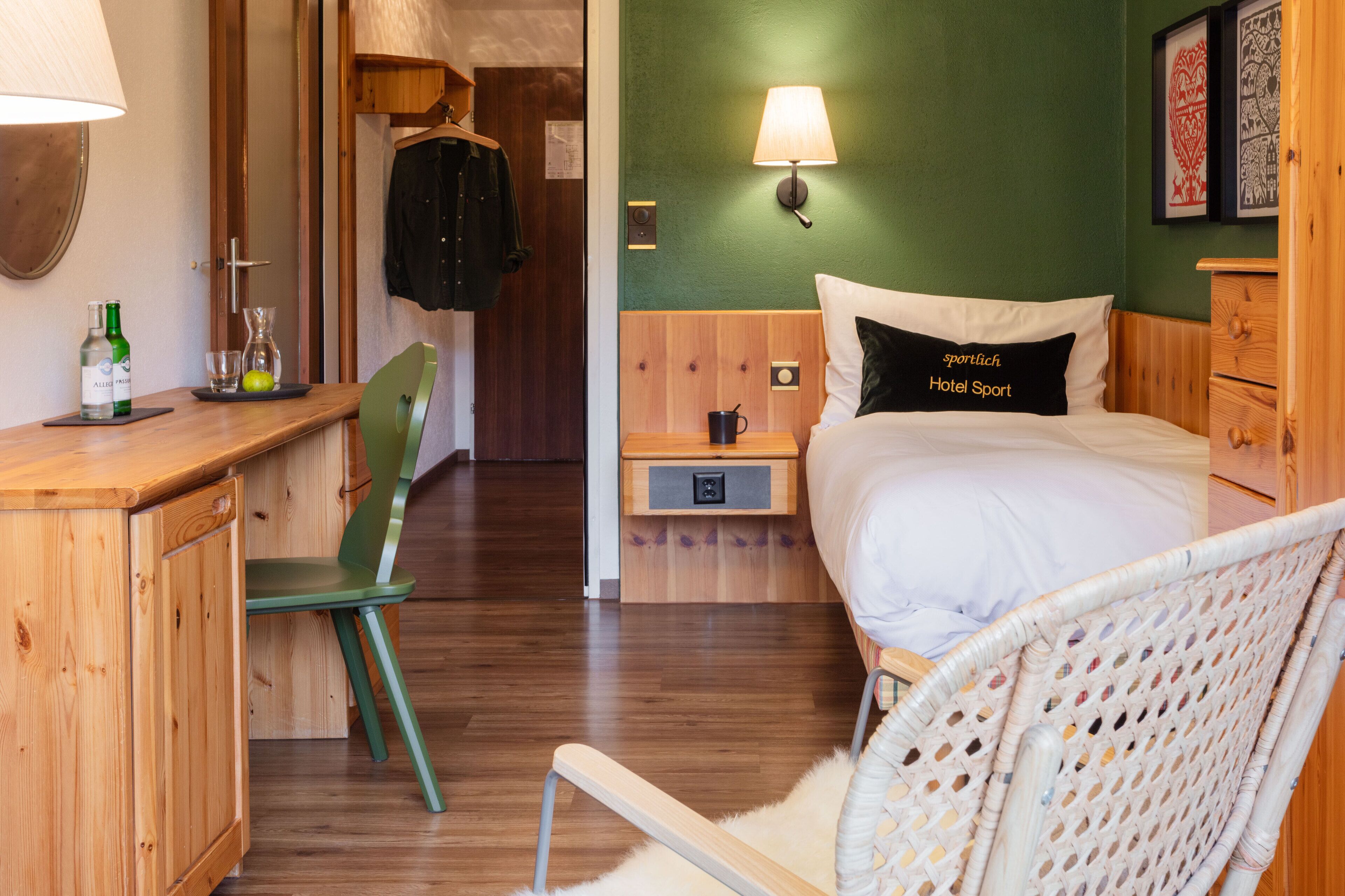 A hotel room with a wooden desk, a green chair, a bed with a pillow labeled "Hotel Sport," a wall-mounted lamp, and a small bedside table with a cup and a glass.