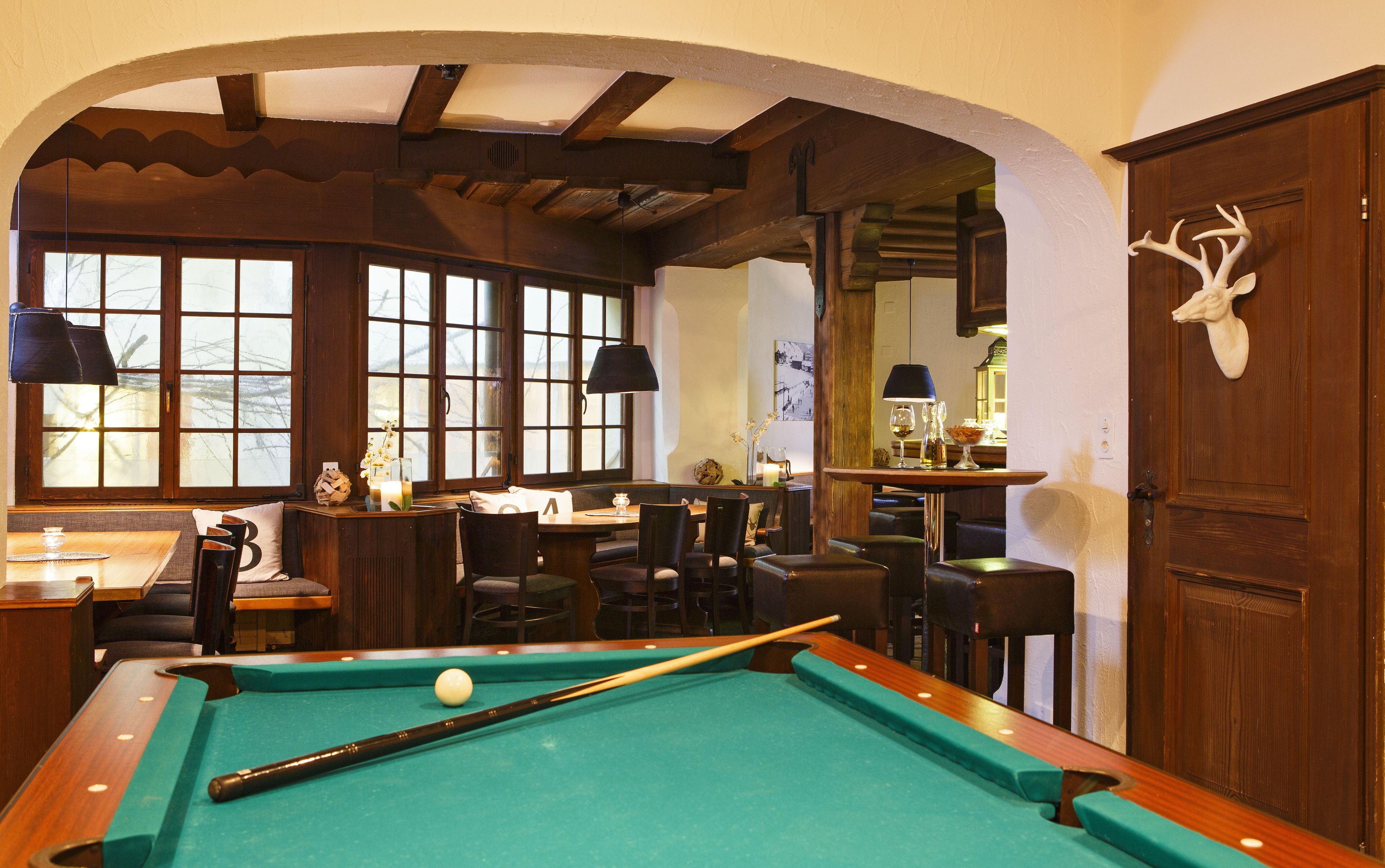Une table de billard dans une pièce meublée de meubles en bois, une tête de cerf accrochée au mur et de grandes fenêtres.