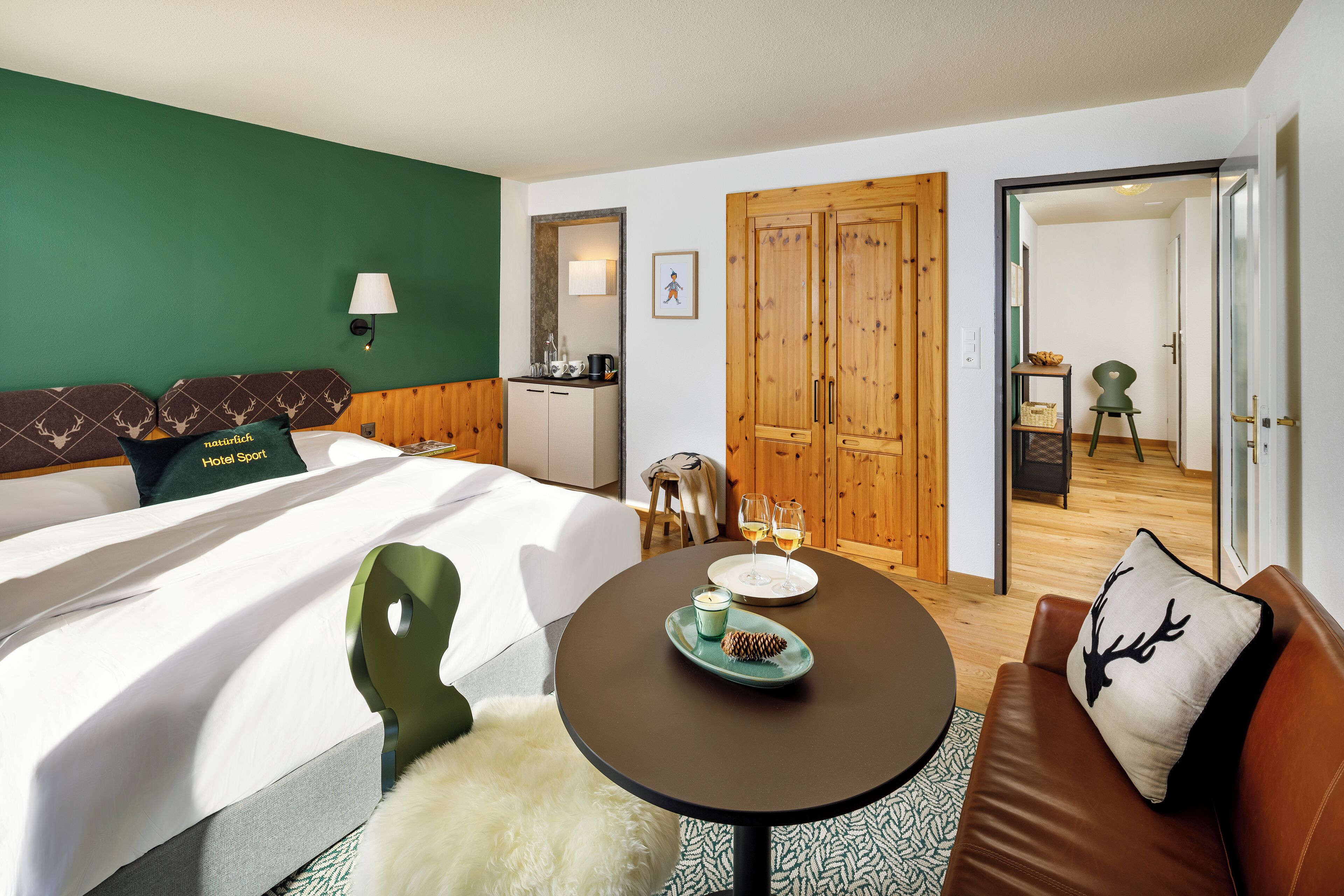 A hotel room with a bed, a round table with a plate and a candle, a green chair, a brown sofa, and a wooden wardrobe.