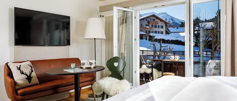 Habitación de hotel con sofá, una mesita con lámpara, una silla y una cama. Hay un televisor montado en la pared y un balcón con vistas a una montaña nevada.