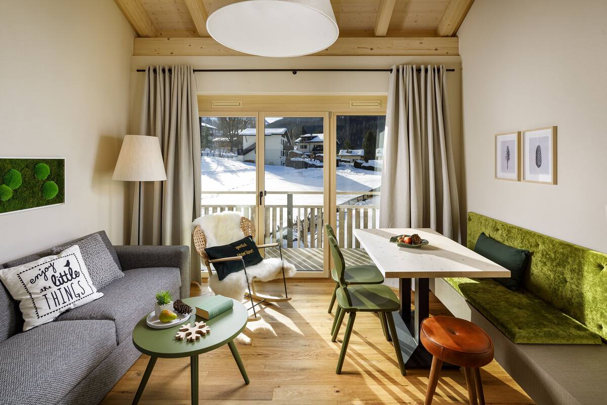 A cozy living room with a sofa, a green couch, a dining table, and a view of a snowy landscape through the glass door.