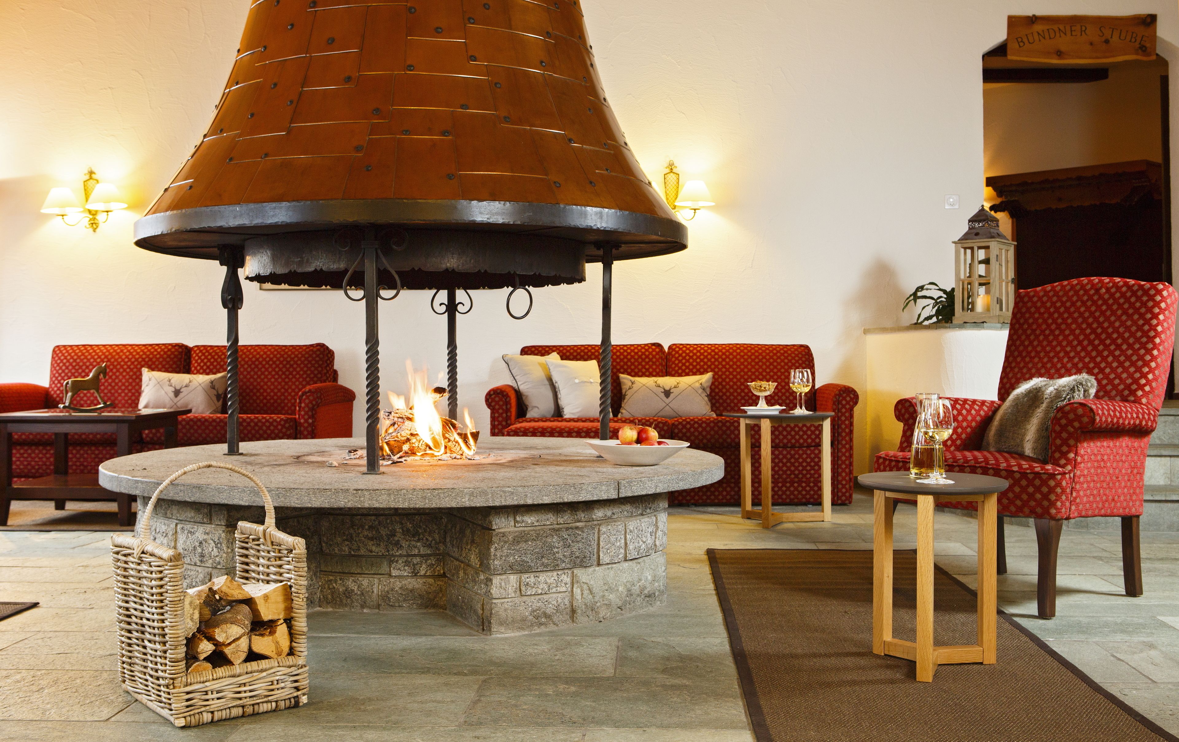 Un rincón acogedor con una chimenea de piedra central, sofás rojos con estampado y una mesita de madera con vasos.