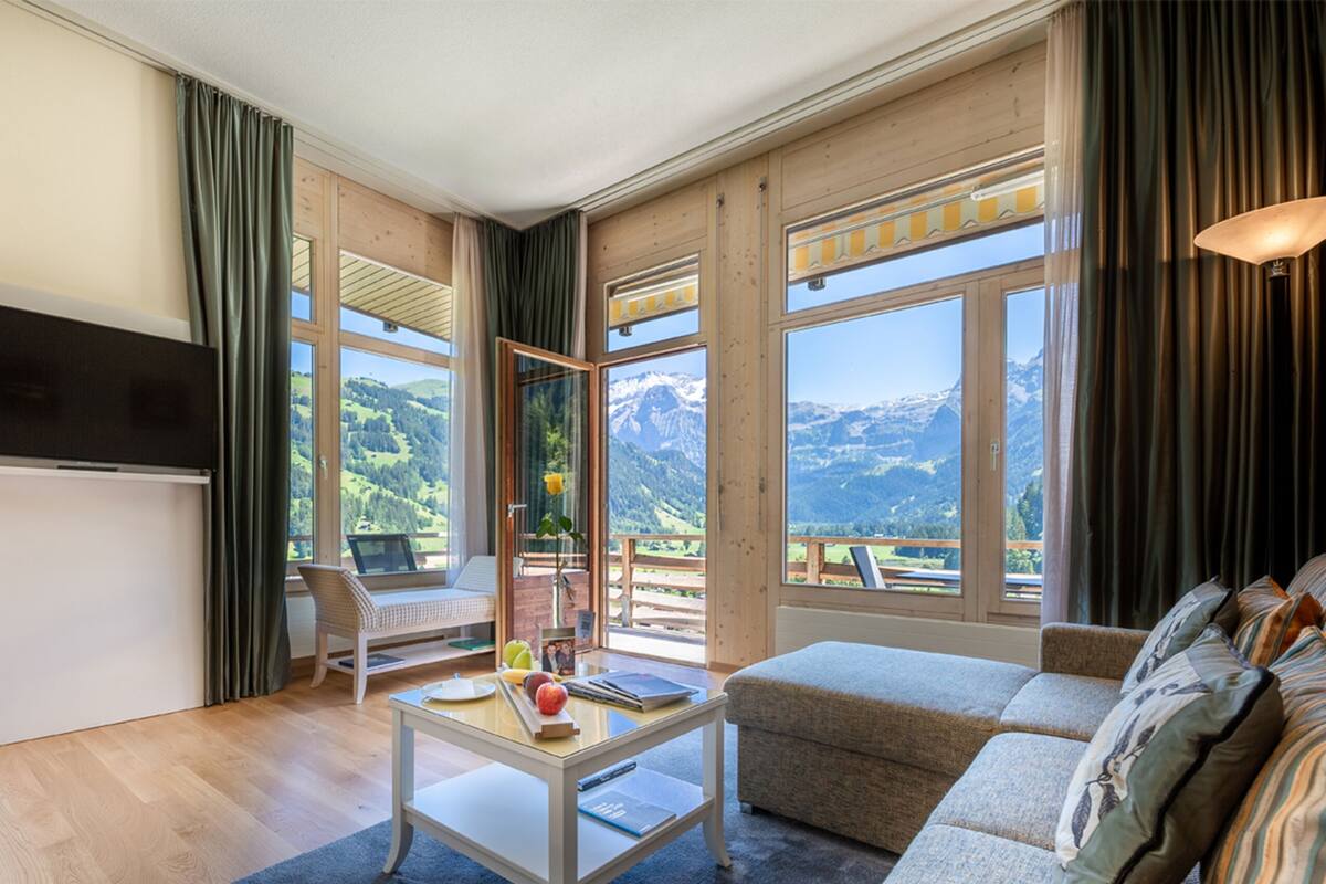 Una habitación con piso de madera, un sofá blanco, una mesita con una laptop, un televisor y ventanas amplias con vista a la montaña.