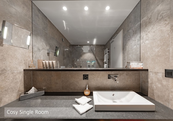 Una habitación individual moderna con un espejo grande, un lavabo blanco y un tocador con artículos de aseo.