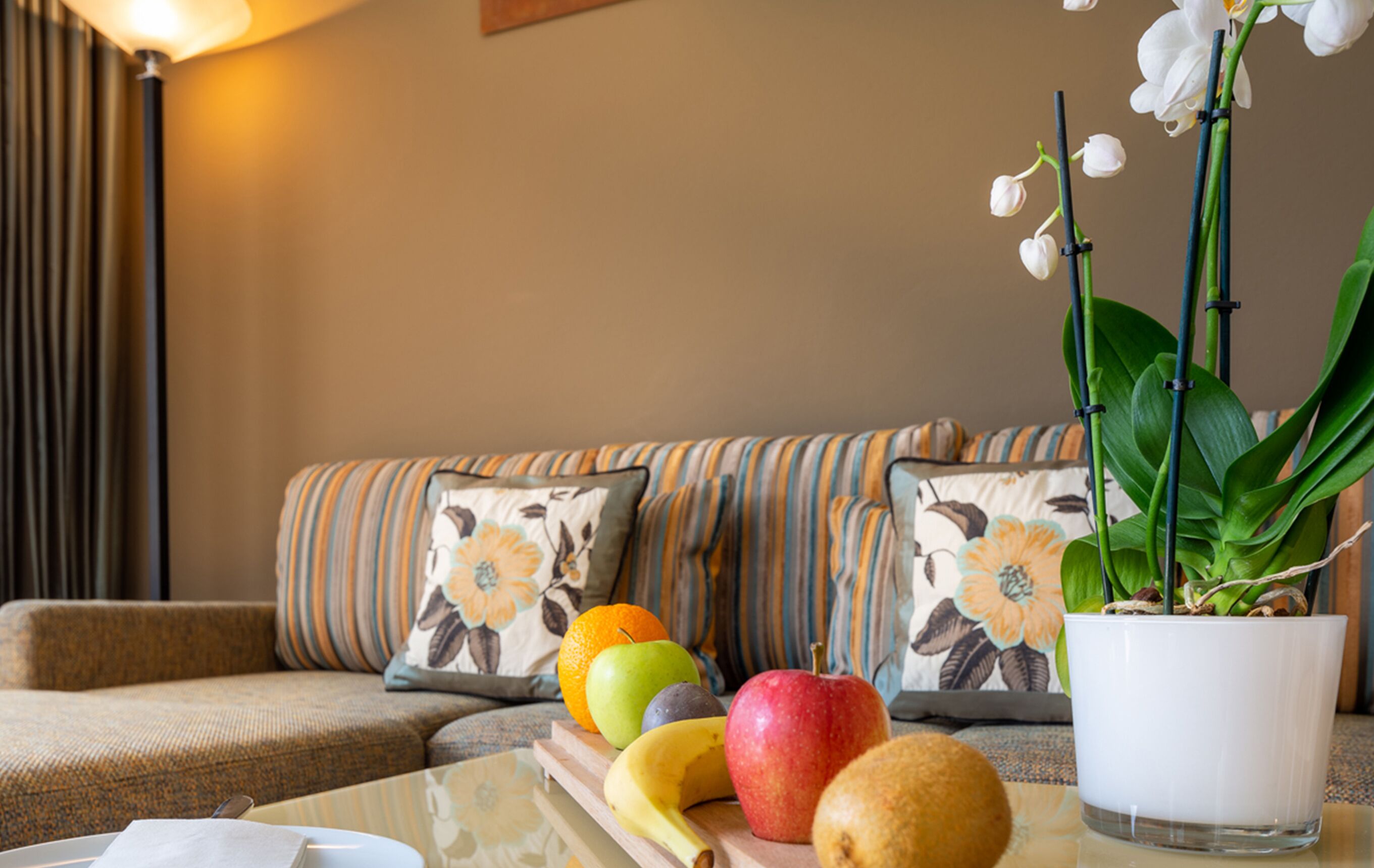A living room with a sofa, pillows, a coffee table with fruits and a potted plant.