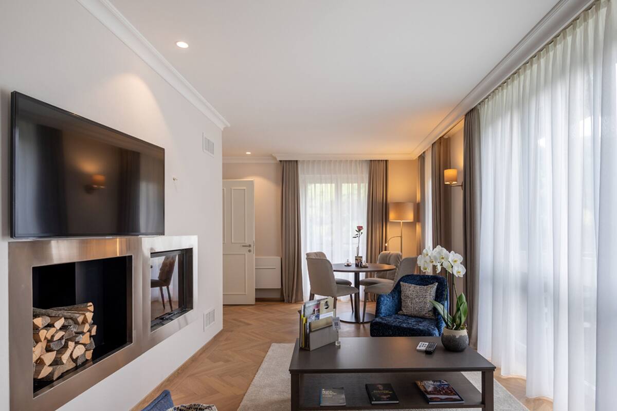 Una habitación de hotel moderna con una televisión de pantalla plana en la pared, una chimenea, una zona de comedor con mesa y sillas, y un sofá con una orquídea en maceta.