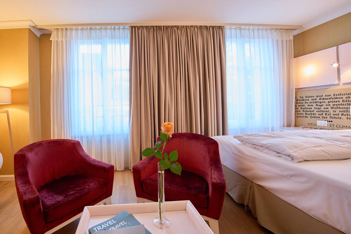 Habitación de hotel con dos sillones rojos, una cama, una mesita de noche con lámpara, un jarrón con una rosa y una ventana con cortinas.