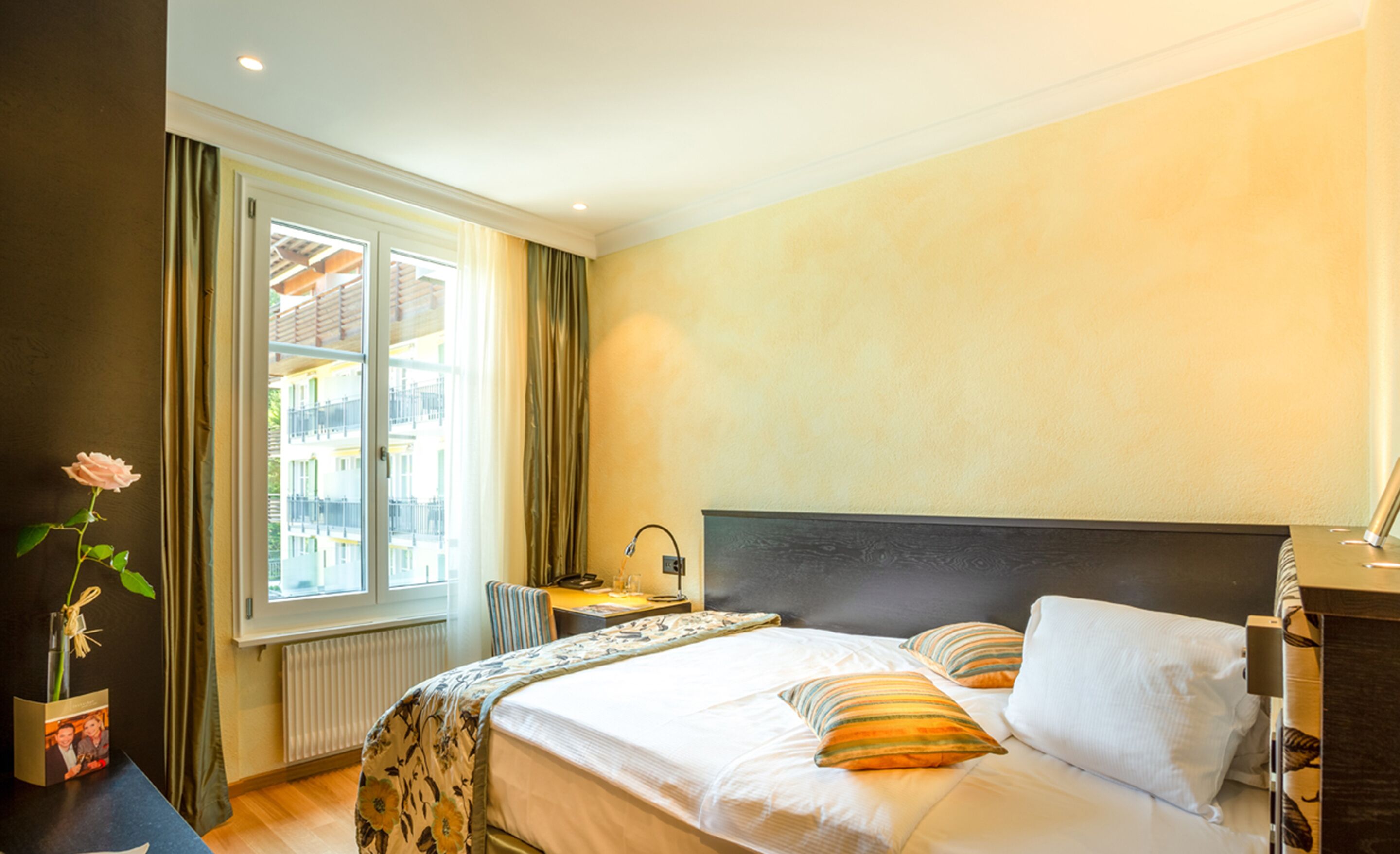 A hotel room with a bed, a desk, a chair, a window with curtains, and a vase with a flower.