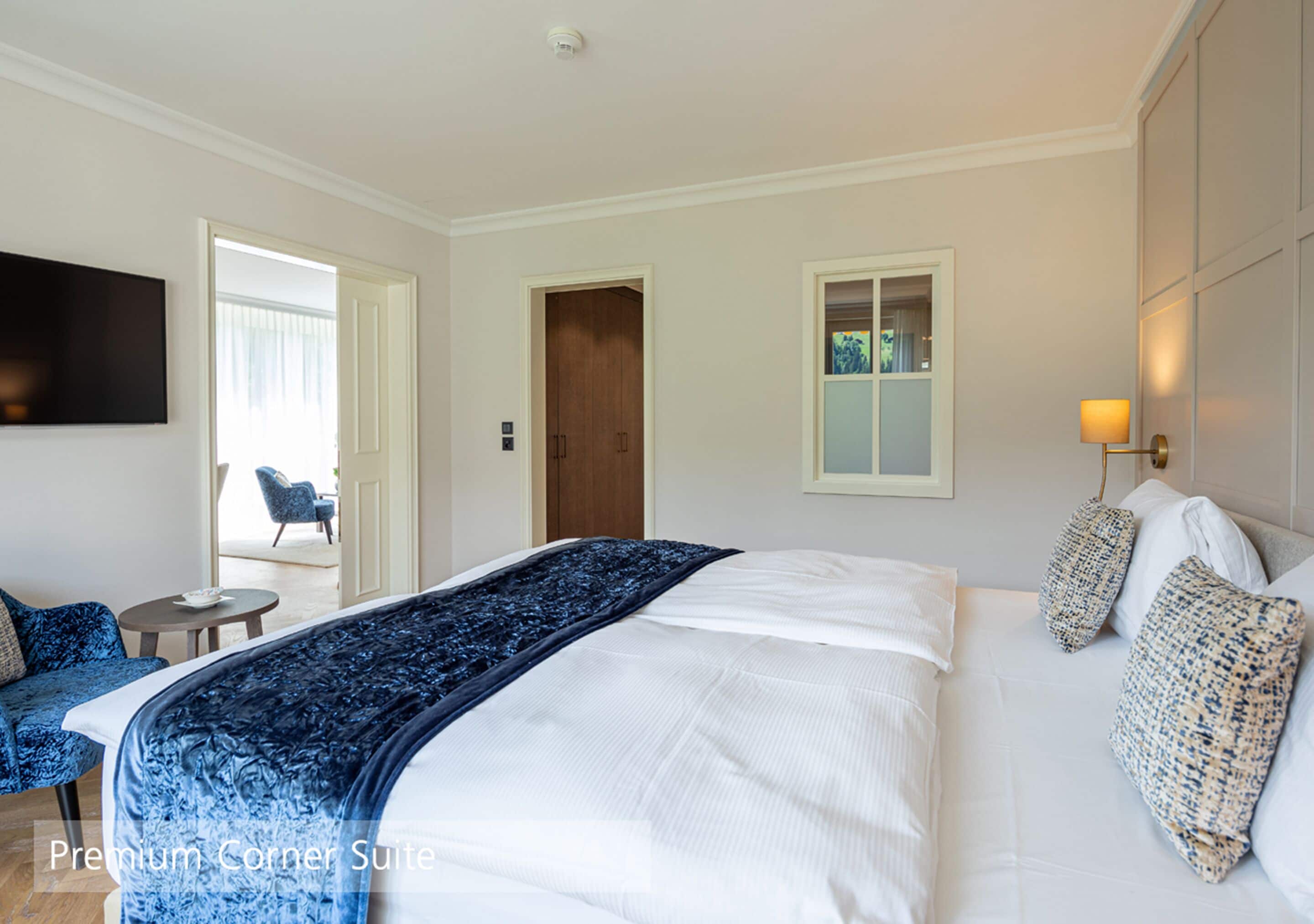 Une chambre d’hôtel avec un grand lit, une télévision, une petite table et une chaise. Il y a une porte qui donne sur une autre pièce et une fenêtre avec des rideaux.