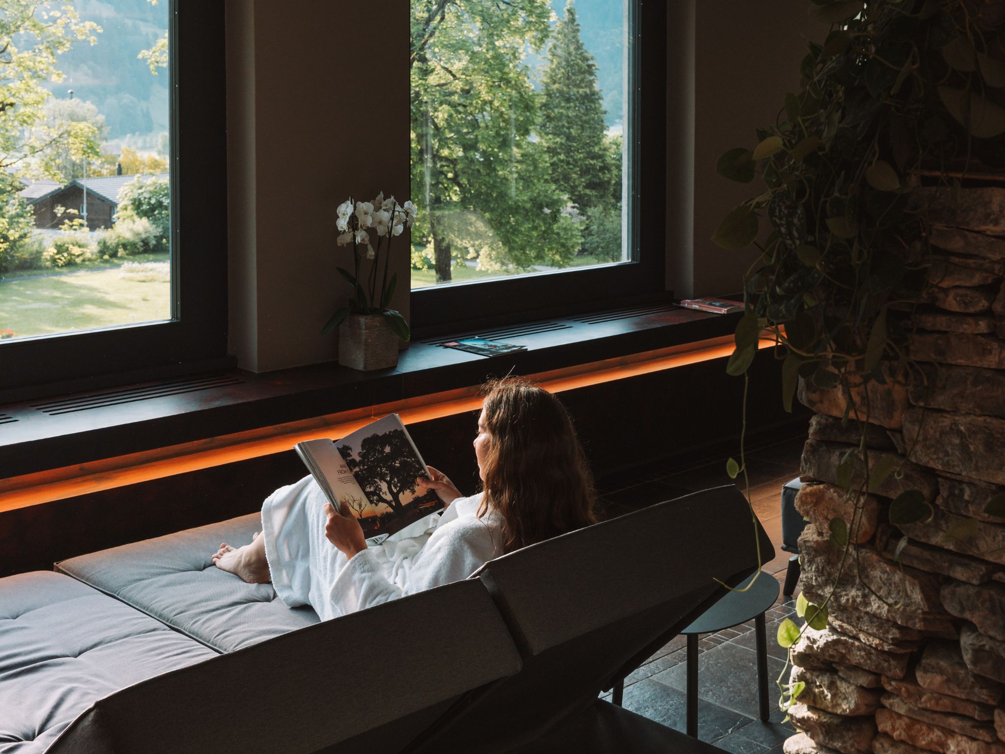 A person reading a book in a room with a large window, a bench, and a stone wall.
