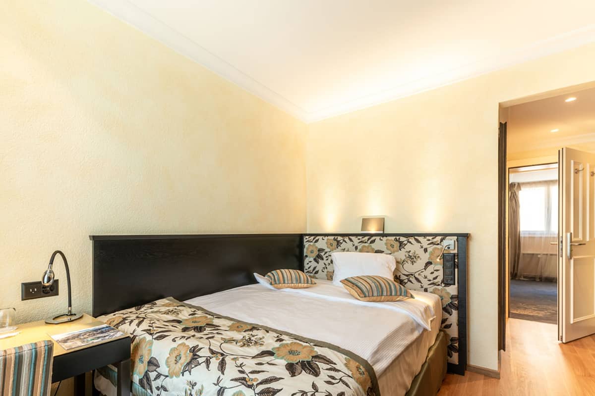 Habitación de hotel con cama, mesita de noche, lámpara y vista al pasillo.