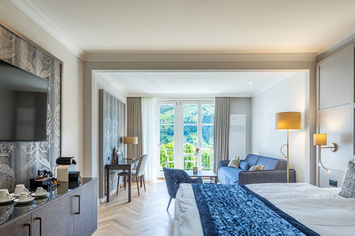 Habitación de hotel con cama, sofá, escritorio con silla, televisor y ventana grande con vistas a zonas verdes.