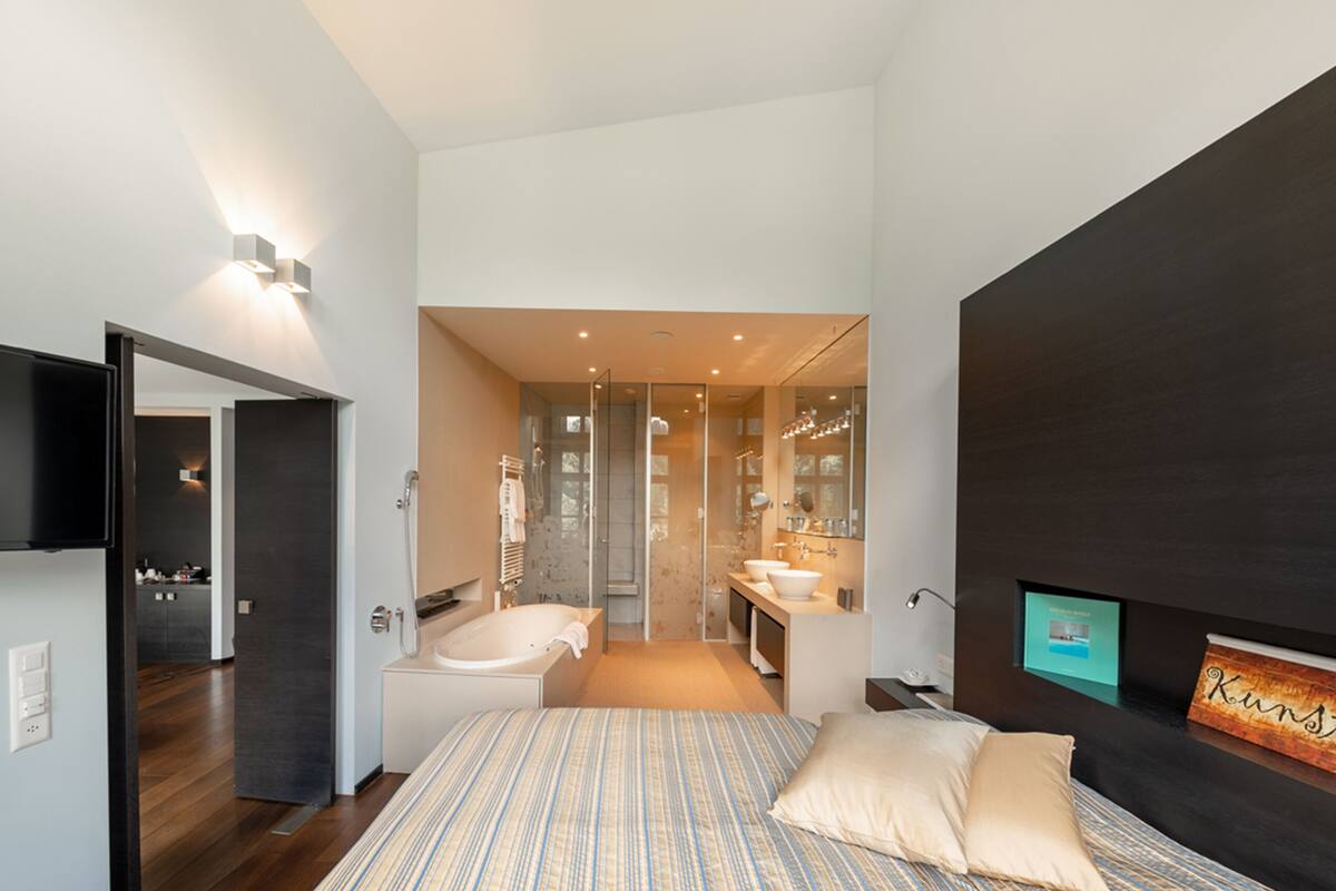 Un dormitorio moderno con una cama grande, un televisor, una pared de acento oscura y un baño visible a través de una puerta abierta.