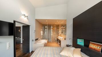 Une chambre moderne avec un grand lit, une télévision, un mur d’accent de couleur sombre et une salle de bain visible à travers une porte ouverte.