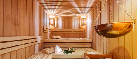 A wooden sauna with benches, a hanging copper basin, and wall-mounted lights.
