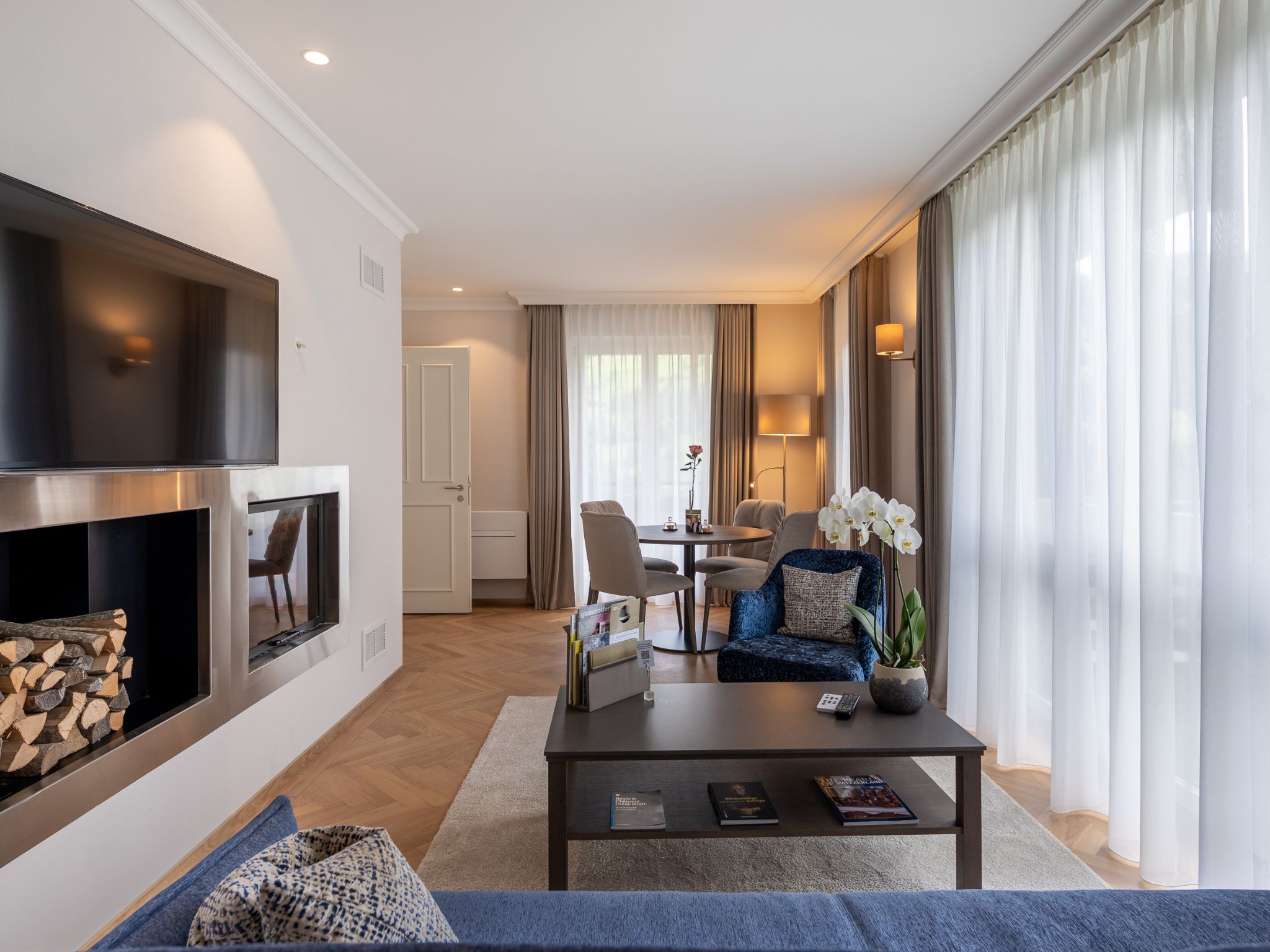 A modern living room with a fireplace, a dining area, and a seating area with a blue sofa and orchid plant.