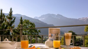 Una mesa de desayuno con vista a la montaña, que incluye una tetera, tazas y un vaso de zumo de naranja.