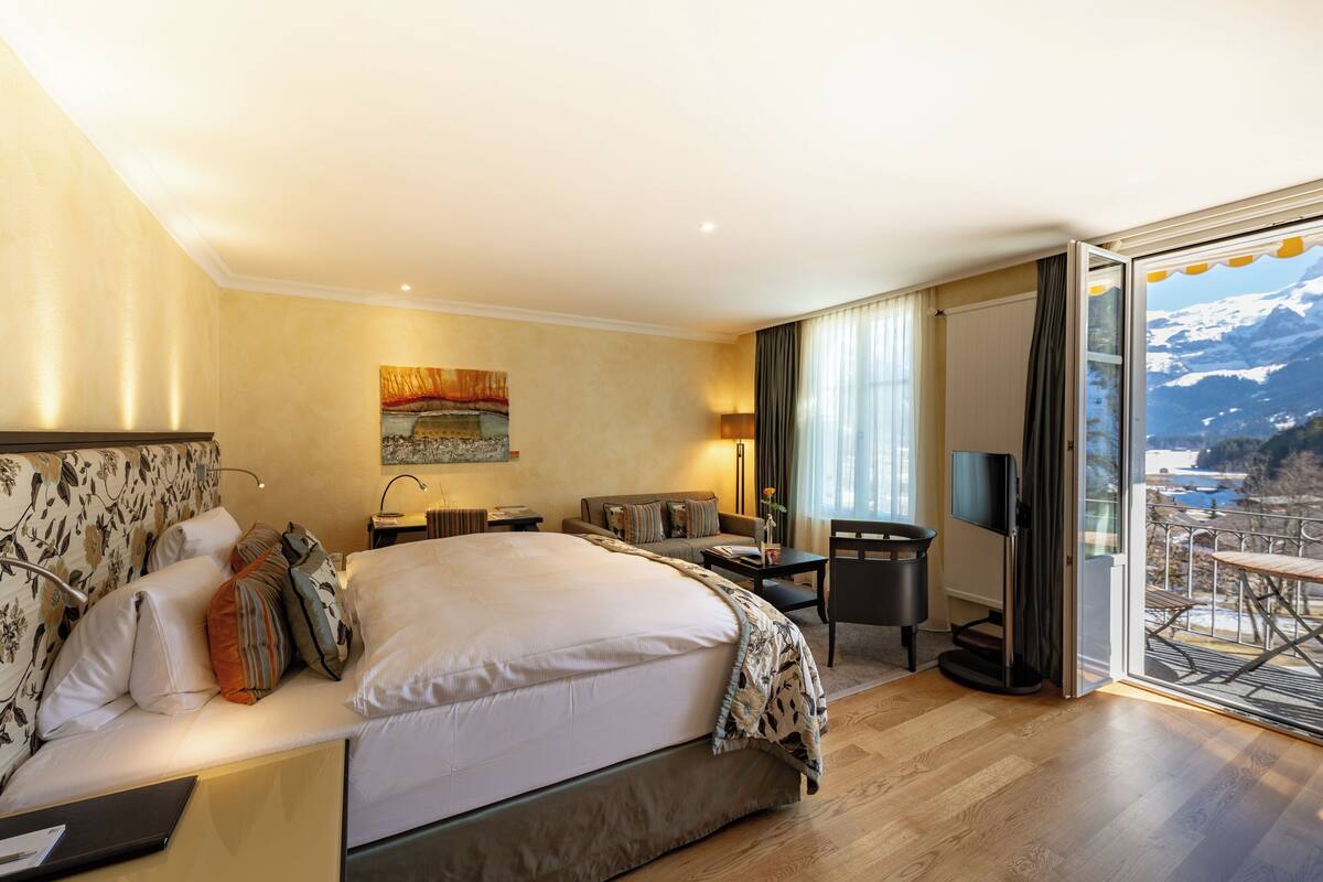 Habitación de hotel con cama, escritorio, silla, televisor y balcón con vista a la montaña.