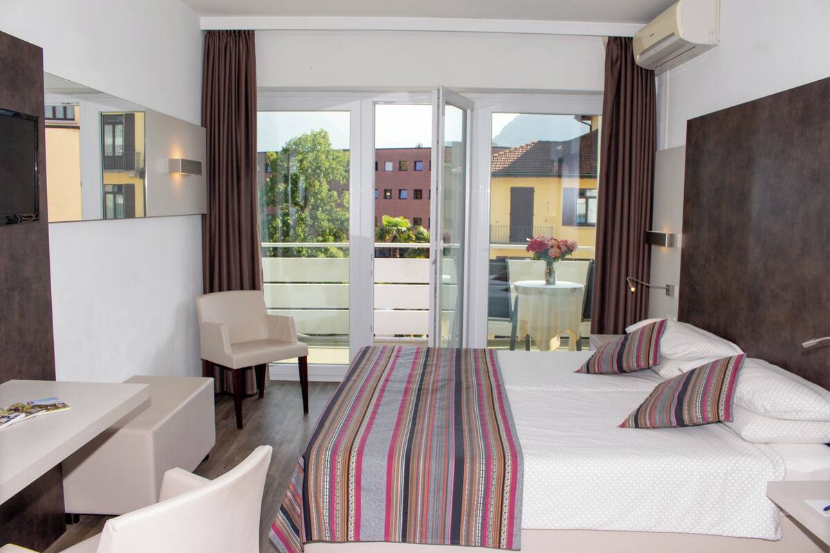Ett hotellrum med en säng, ett skrivbord, en stol och en balkong med utsikt över byggnader och träd.