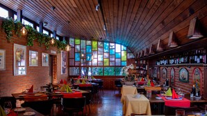 Un restaurante con paredes de ladrillo, techo de madera y ventanas amplias con vidrieras de colores. Las mesas están dispuestas con servilletas y platos rojos.